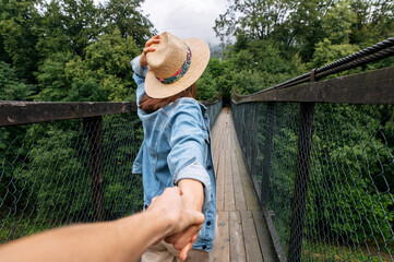A girl in a hat is walking on suspension bridge and leading someone by hand. Follow me photo. Back view - obrazy, fototapety, plakaty