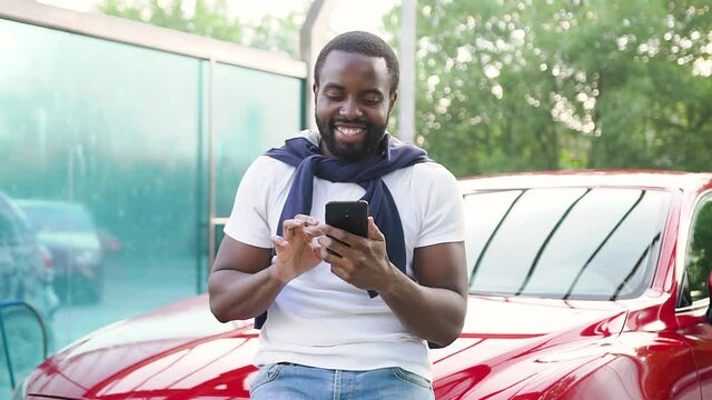 Handsome smiling bearded young black-skinned guy standing near his own red expensive car in car wash and enjoying revision funny pictures on phone