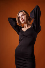 charming elegant fashion model wearing black dress with deep neckline posing on orange background. asian skinny young woman sits in sexy evening gown. beautiful sensual glamorous girl poses in studio