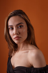 charming elegant fashion model wearing black dress with deep neckline posing on orange background. asian skinny young woman sits in sexy evening gown. beautiful sensual glamorous girl poses in studio