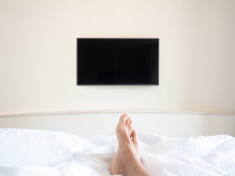 Blank Television On White Wall In Front Of The Bed. First Person View From Bed Seeing Men Feet And TV.
