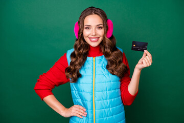 Photo of pretty lovely young lady smiling showing credit card hand hip bank promotion profitable deposit saving wear pink ear warmers blue vest red sweater isolated green color background