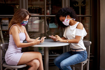 Two women in stylish masks sitting in a cafe and holding their mobile phones