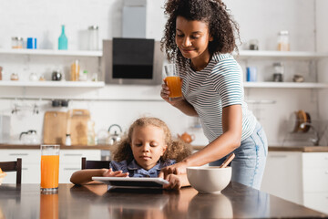 african american mother holding orange juice and touching digital tablet near concentrated child in kitchen