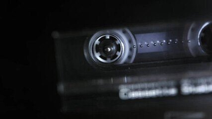 An audio cassette spools tape in extreme macro. Shallow depth of field with focus on the rorating spool. Suggests an interrogation or phonetap. 