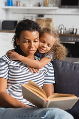 selective focus of african american girl embracing nanny in striped t-shirt reading book on couch