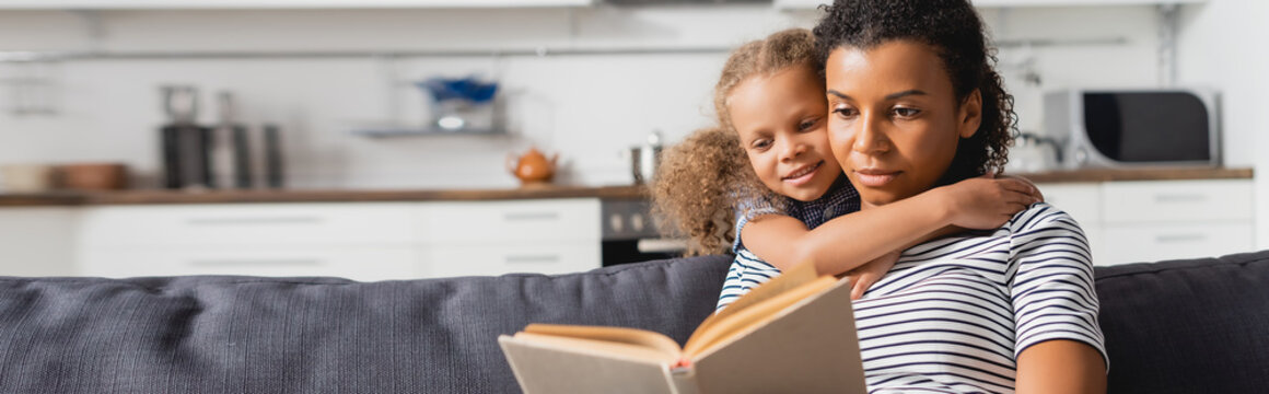 horizontal image of african american girl embracing nanny sitting on couch and reading book in kitchen