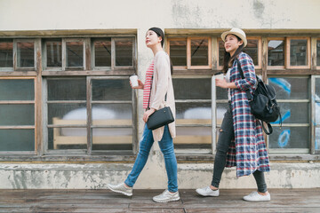 Young asian female people walking around old city. full length two stylish girl friends having fun sightseeing in little village. two women tourists hold coffee cpus pass window of historical house