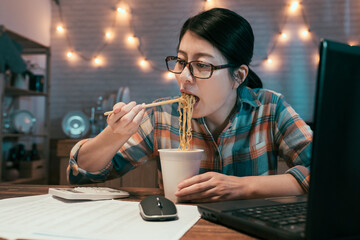 elegant businesswoman eating instant noodles at dining table while over work on project with laptop computer and calculator. hungry asian korean female in glasses having bedtime snacks in night.