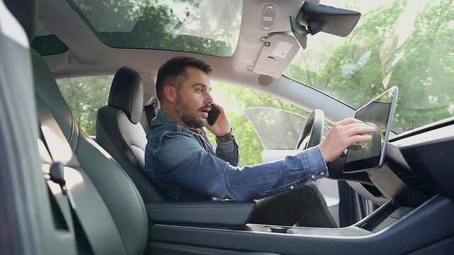 Attractive concentrated bearded modern man sitting at helm of his expensive car,talking on mobile and using gps navigator,close up