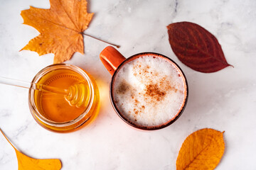 Top view of coffee with milk foam and cinnamon, jar of honey and orange autumn leaves