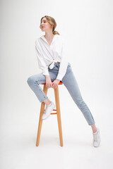 portrait of young pensive caucasian woman posing in shirt and blue jeans, sitting on stool in white studio. model tests of pretty girl in basic clothes on cyclorama. attractive, charming female poses