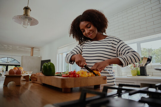 Happy african woman chopping yellow and red bell peppers on wooden chopping board in modern kitchen