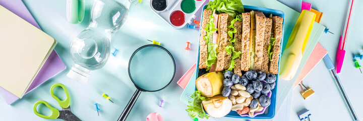 Healthy children school lunch box: sandwich, vegetables ,fruit and mineral water bottle, with school supplies on, on light blue background copy space