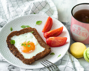 One baked chicken egg in a slice of bread. On a white plate, next to pieces of tomato. Sprinkled with basel leaves .. A mug of tea and sweet macaroon cookies. Hearty breakfast. Homemade food.
