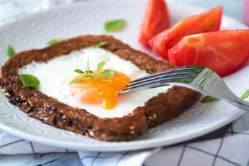 One baked chicken egg in a slice of bread. On a white plate, next to pieces of tomato. Sprinkled with basel leaves. The fork pierces the yolk, it flows out. Hearty breakfast. Egg dishes.