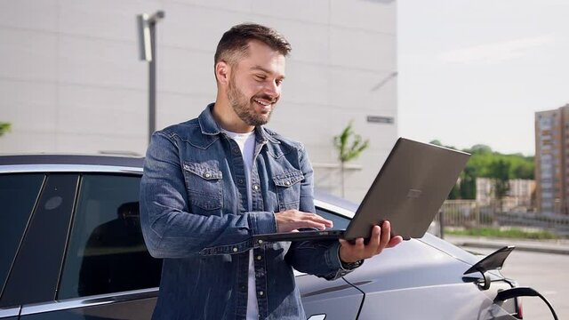 Good-looking smiling young bearded man has video call on computer while waiting when charging his luxurious electocar in charging station