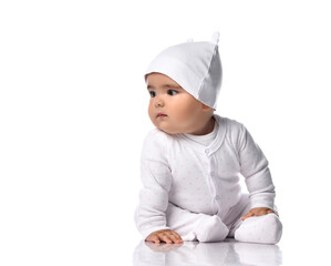 Curious infant baby toddler sits on the floor in white onepiece jumpsuit overall and funny hat with ears and looks aside