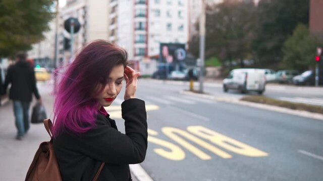 Pretty asian hipster girl waiting at a bus stop on a busy street. Woman tourist at sidewalk. Concept of lifestyle, travel and tourism.Pink colored hair charming millenial using public city transport.