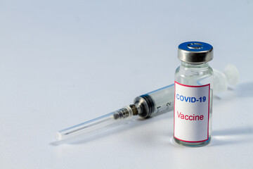 In the foreground is a bottle of COVID-19 vaccine. In the background is a medical injection syringe.
