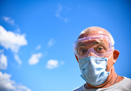 Senior man wearing medical mask and goggles looking at the camera close-up on a blue sky with clouds background. coronavirus concept. respiratory protection