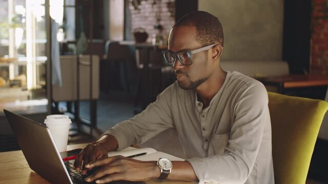 Young African American businessman in casualwear sitting at table in cafe with disposable coffee cup and notepad and typing on laptop while working on break