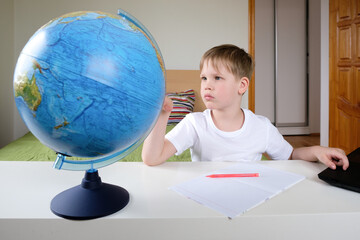 Distance learning, geography lesson. The boy is sitting on the bed with a globe at the laptop.