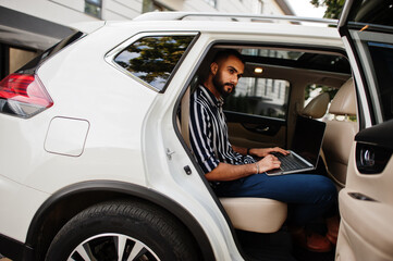 Successful arab man wear in striped shirt and sunglasses pose inside white suv car with laptop in hands.