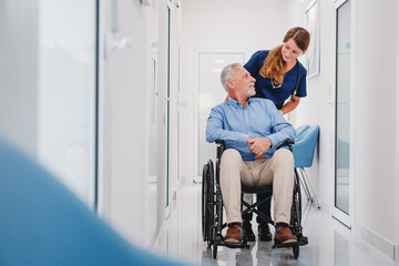 Senior male patient sitting in wheelchair in hospital corridor with female nurse doctor - Powered by Adobe
