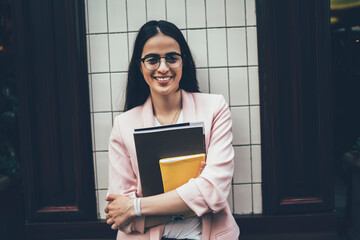 Half length portrait of cheerful female journalist with sketchbook and notepad posing at urban setting, successful woman in classic glasses for eyes correction smiling at camera during leisure in city
