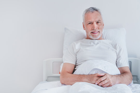 Portrait Of Male Senior Patient Lying In Hospital Bed Smiling At Camera