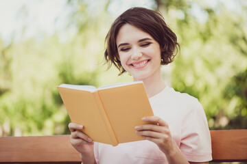 Fototapeta premium Photo of attractive sweet young lady toothy smile sit bench hold read yellow book diary interesting story spend free time hobby learn new information wear pink t-shirt park outdoors