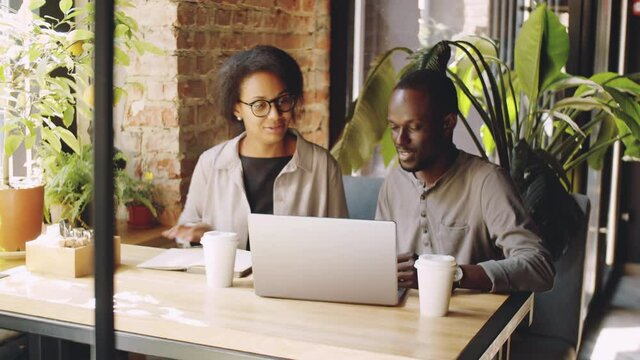 African American male and female colleagues sitting at cafe table with disposable coffee cups and discussing business project on laptop