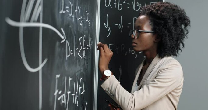 African American young woman teacher at school writing formulas and mathematics laws on blackboard. School concept. Female math lecturer in glasses explaining physics laws. Education.