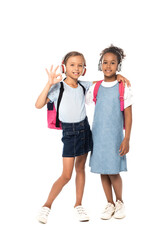 schoolkid listening music in wireless headphones, hugging african american friend and showing ok sign isolated on white