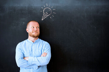 Pensive young businessman standing over chalkboard background with light bulb.