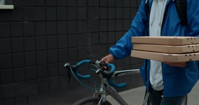 Crop view of male courier carrying carton packaging pizza boxes and pushing bike. Person with backpack delivering order to customer. Concept of delivery service