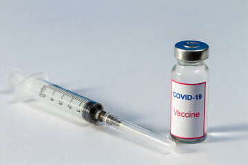 On the right is a bottle of COVID-19 vaccine. On the left is a medical injection syringe. The background is light.