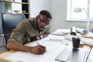 Afro-American architect working in office with blueprints.Engineer inspect architectural plan, sketching a construction project. Portrait of black handsome man sitting at workplace. Business concept. - Powered by Adobe