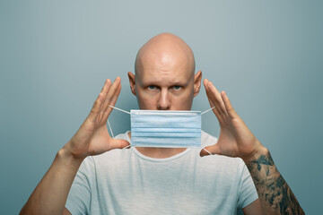 A young handsome bald guy with a tattoo on his arm puts on a protective mask. - 375180622