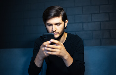 Portrait of young man typing on the smartphone. Sitting on sofa, on the background of black brick wall.