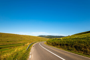 Asphalt road leading through green hills in Transylvania, Romania, blue sky background at early autumn.
