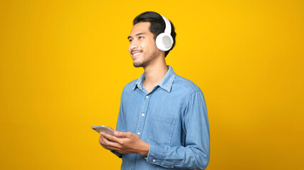 Asian man with headphones listening music using smartphone app while standing over isolated yellow...