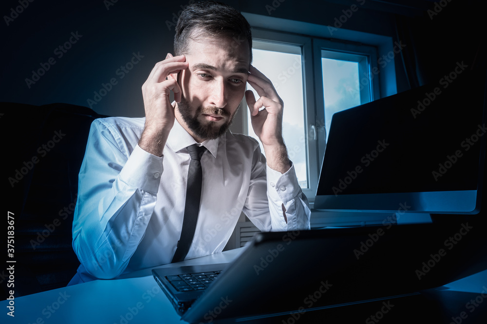 Poster a man sits at a table with a laptop and computer and holds his head. irregular working hours. fatigu