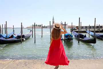 Fototapeta na wymiar Holidays in Venice. Back view of beautiful girl in long red dress enjoying view of Venice Lagoon with the island of San Giorgio Maggiore and gondolas moored.
