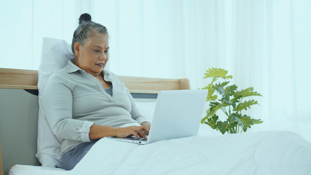 Happy Indian Elderly Woman Sitting On Her Bed Using A Laptop. Online Shopping On The Website. Patient At Home By Tele Health Using The Laptop In Treatment Of Diseases. Virtual Doctor Appointment