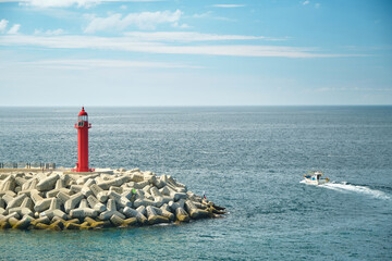 Blue sky and sea. A fishing boat passing by a breakwater with a red lighthouse.