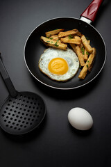 fried egg in black pan with french fries and parsley, on black background and slotted spoon and raw egg