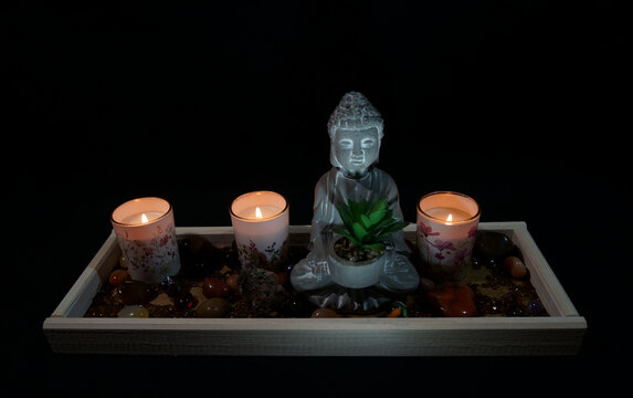 Statue eines meditierenden Buddha mit drei brennenden Kerzen und einer Pflanze vor einem schwarzen Hintergrund