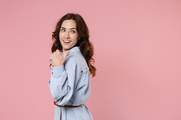 Side view of cheerful attractive funny young brunette woman 20s wearing casual blue shirt dress pointing thumb aside on mock up copy space isolated on pastel pink colour background studio portrait.